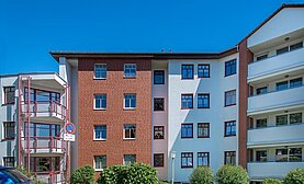 Frontansicht des Mehrfamilienhauses in der Friedrich-List-Straße 18: die Fassade hat weiße Elemente und Mauern aus rotem Backstein und breite Balkone. Die Einfahrt ist umgeben von Büschen. 